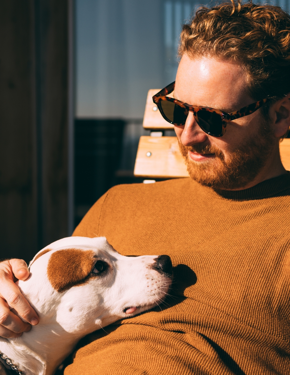 Mann mit Sonnenbrille auf einer Liege mit Hund auf dem Schoss.
